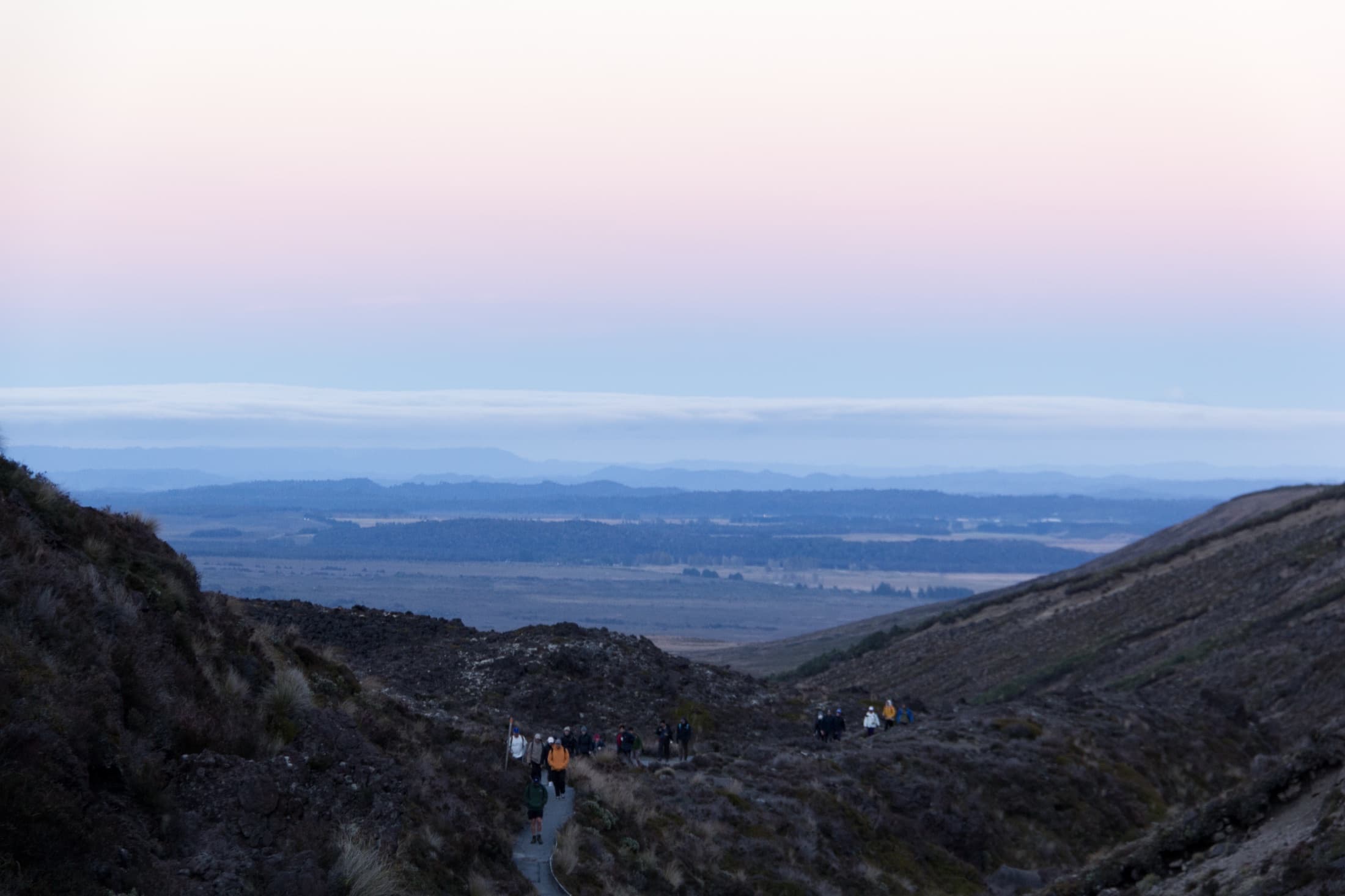 Hikers descending the trail at dawn with a wide valley and pink sky beyond